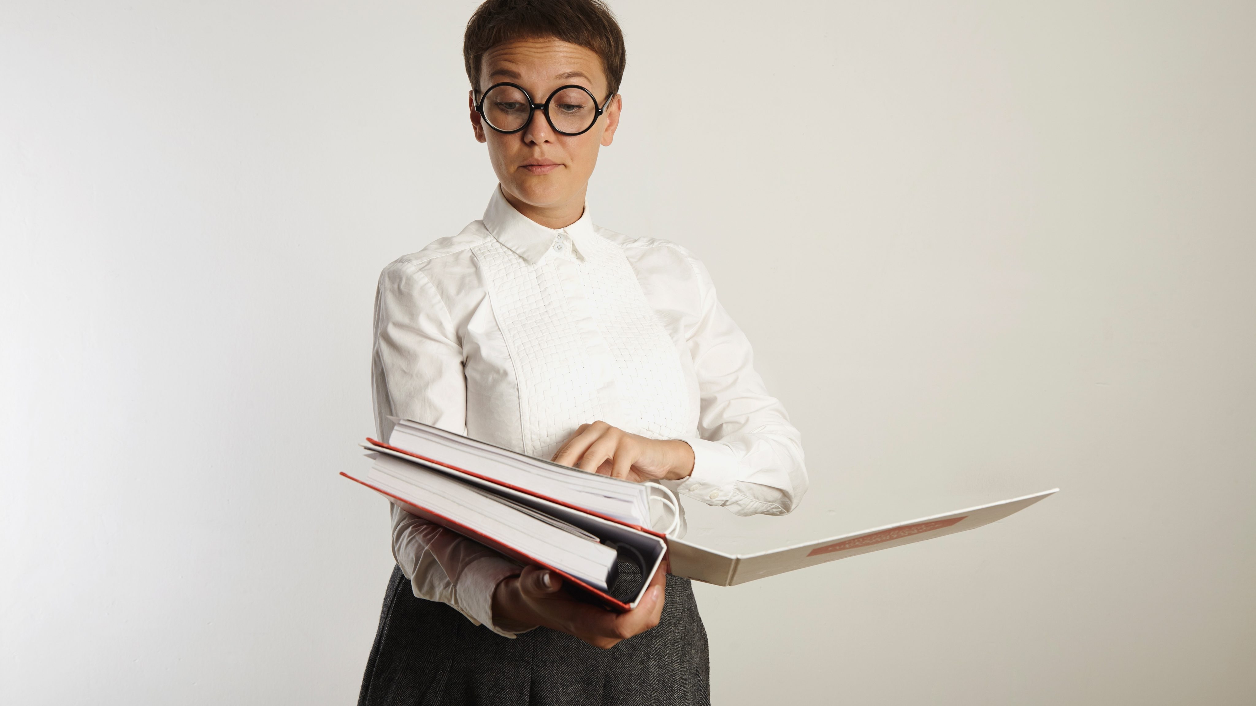 Disapproving female teacher wearing white blouse gray tweed skirt and glasses grading papers in heavy binders isolated on white