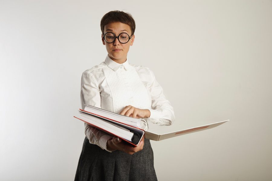 Disapproving female teacher wearing white blouse gray tweed skirt and glasses grading papers in heavy binders isolated on white