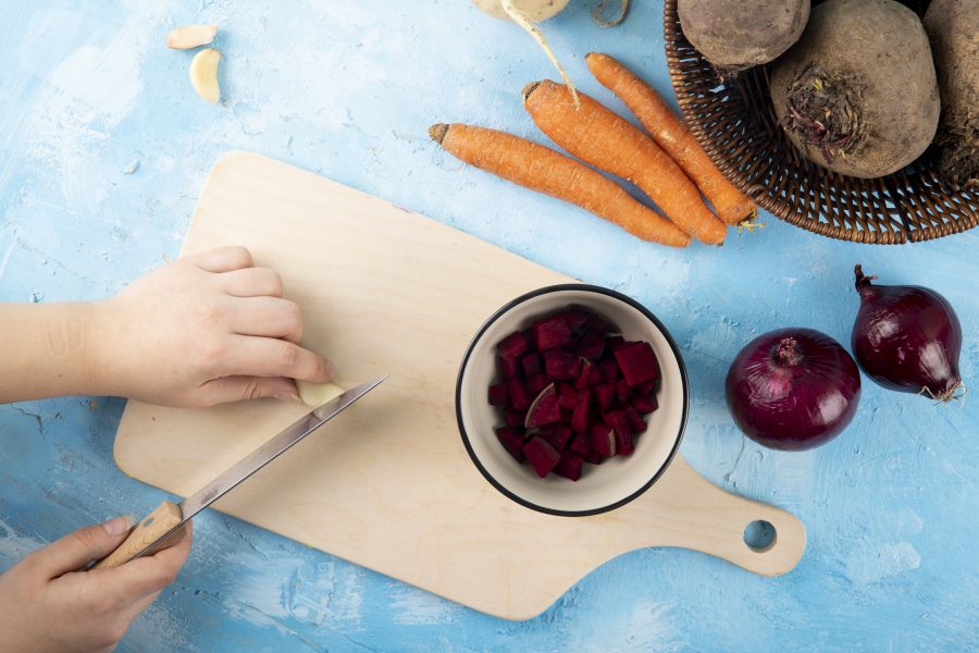 Top view woman cuts garlic on board with chopped beets in cup with carrots and red onions on table