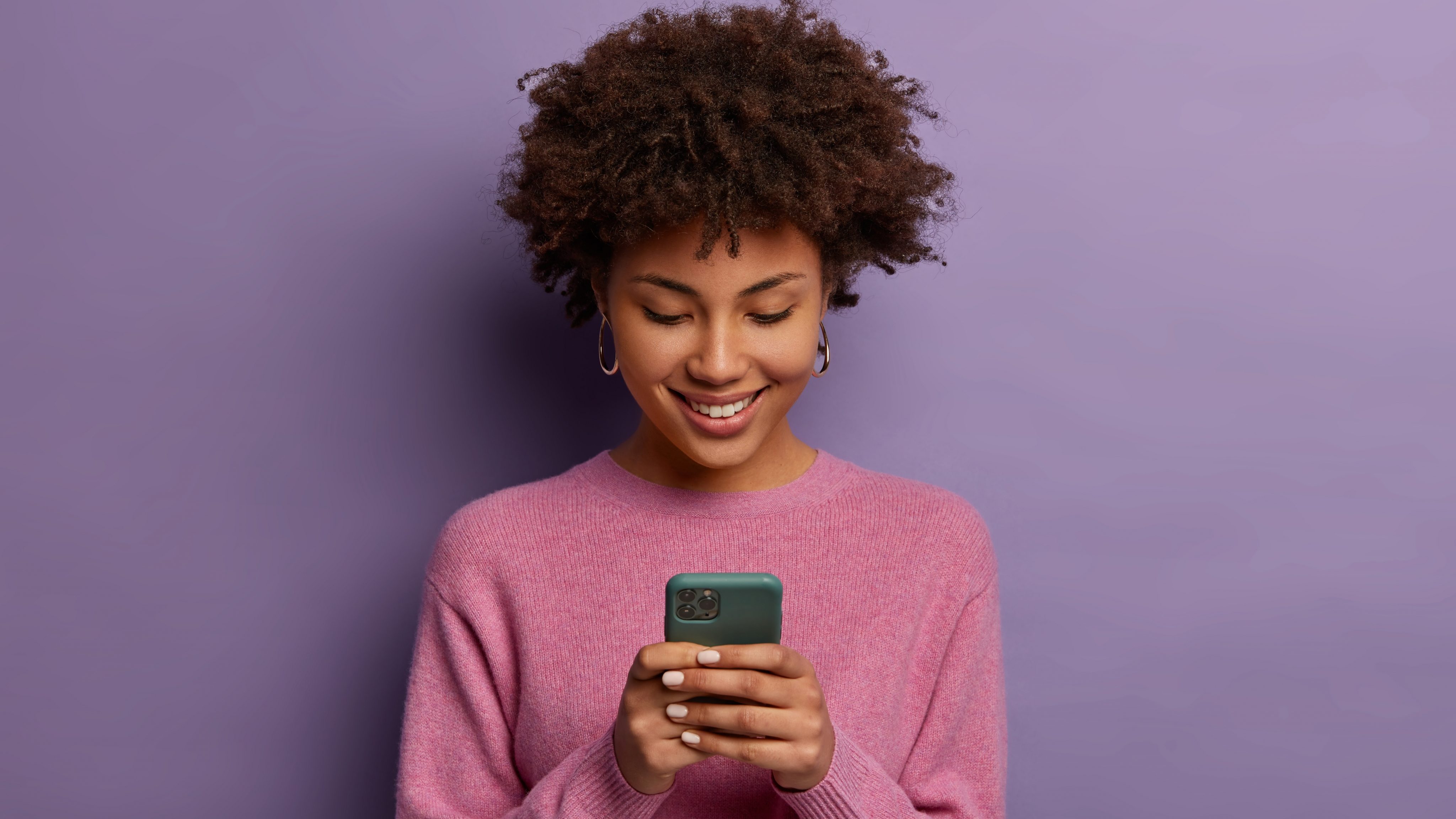 Pretty curly woman holds modern mobile phone types messages on smartphone device enjoys online communication downloads special app for chatting smiles tenderly isolated on purple wall