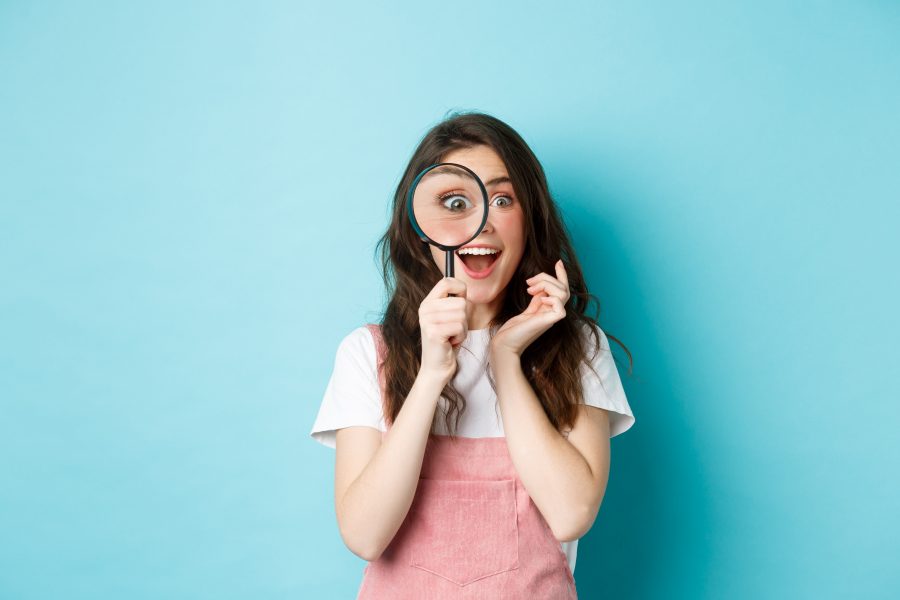 Happy young woman looking through magnifying glass with excited face found search something standing blue background